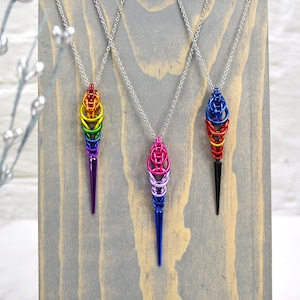 May include: Three colorful metal chainmaille pendants hanging on a wooden display stand. The pendants are shaped like ice cream cones and are in rainbow, pink and blue, and black colors.