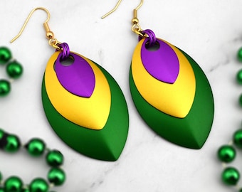May include: A pair of gold-tone earrings with a layered design in green, gold, and purple. The earrings are in a teardrop shape and have a hook closure. Green beaded necklaces are in the background.