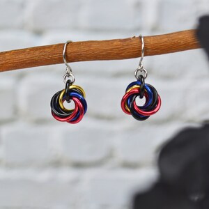 May include: A pair of handmade earrings with a colorful chainmaille design. Each earring features a cluster of interlocking rings in red, black, blue, and gold, suspended from a silver hook. The earrings are displayed against a blurred background.