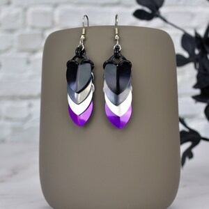Puede incluir: Un par de pendientes colgantes con un esquema de color negro, blanco y morado. Los pendientes están hechos de metal y tienen un diseño en forma de corazón.
