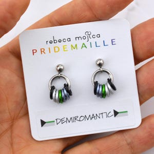 May include: A pair of silver hoop earrings with a ring of green, gray, and black beads. The earrings are on a white card with the text "rebeca mojica PRIDEMAILLE" and "DEMIROMANTIC" on it.