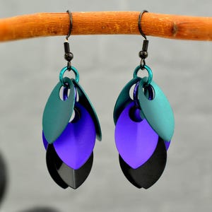 Puede incluir: Un par de pendientes colgantes hechos a mano con un diseño de escamas. Cada pendiente presenta tres escamas metálicas superpuestas en verde azulado, morado y negro. Los pendientes están suspendidos de ganchos negros y anillas turquesas.