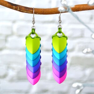 Puede incluir: Un par de pendientes colgantes con un diseño de escamas de dragón verde, azul, morado y rosa. Los pendientes están hechos de arcilla polimérica y tienen ganchos plateados.