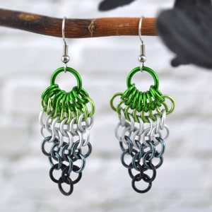May include: A pair of handmade earrings with a chainmail design. The earrings feature a gradient of colors, starting with green at the top, transitioning to silver, then to dark blue, and ending with black at the bottom. The earrings are suspended from silver hooks.