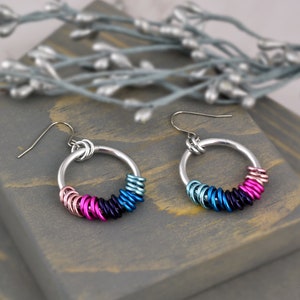 May include: A pair of silver hoop earrings with a gradient of pink, purple, blue, and teal colored rings. The earrings have a simple hook closure.