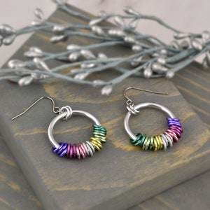 May include: A pair of silver hoop earrings with colorful metal rings. The rings are arranged in a rainbow pattern, with purple, pink, silver, gold, green, and blue rings.