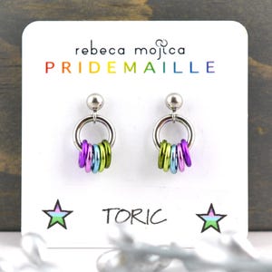 May include: A pair of silver stud earrings with a rainbow of coloured rings hanging from each. The earrings are displayed on a white card with the text "rebeca mojica PRIDEMAILLE TORIC" and two stars.