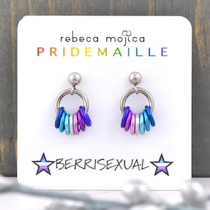 May include: A pair of silver hoop earrings with a ring of colourful beads in blue, purple, pink, and white. The earrings are on a white card with the text "rebecca mojica PRIDEMAILLE" and "BERRISEXUAL" with a star and a blue, pink, and white stripe.