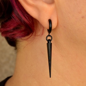 Puede incluir: Pendientes de aro negros con una larga y afilada espiga negra que cuelga de la parte inferior.