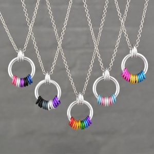 May include: A collection of silver necklaces, each featuring a circular pendant adorned with colorful rings. The rings are in various color combinations, including rainbow, pink, blue, and black. The necklaces hang against a gray backdrop.