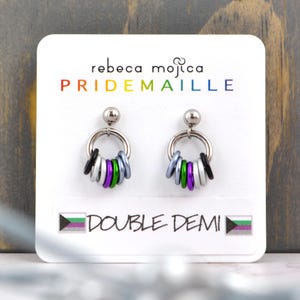 May include: A pair of silver hoop earrings with a ring of colorful beads in the colors of the genderqueer pride flag. The earrings are displayed on a white card with the text "rebeca mojica PRIDEMAILLE DOUBLE DEMI" and a genderqueer pride flag.