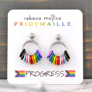 May include: A pair of silver hoop earrings with colourful rings representing the progress pride flag. The earrings are on a white card with the text "rebeca mojica PRIDEMAILLE PROGRESS" printed on it.