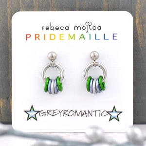 May include: A pair of silver hoop earrings with a dangling charm of green and white rings. The earrings are displayed on a white card with the text "rebeca mojica PRIDEMAILLE GREYROMANTIC".