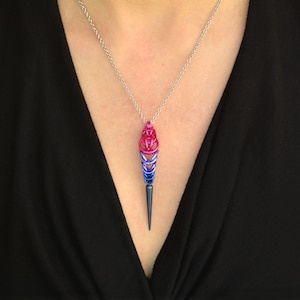 May include: A silver chain necklace with a pendant shaped like a teardrop. The pendant is made of metal and is colored in shades of pink, blue, and purple. The pendant has a pointed tip.