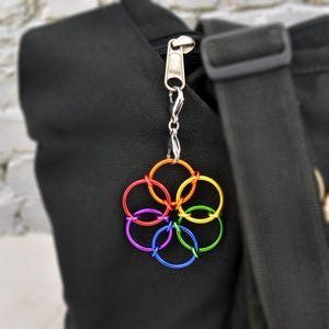 Puede incluir: Un colorido dije en forma de flor con seis anillos entrelazados en tonos arcoíris: rojo, naranja, amarillo, verde, azul y morado. El dije está sujeto a una cadena plateada y a un tirador de cremallera, colgando de una bolsa negra.