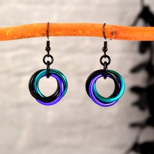 Puede incluir: Un par de pendientes hechos a mano con un diseño de cota de malla. Cada pendiente tiene tres anillos entrelazados en negro, turquesa y morado. Los pendientes cuelgan de ganchos negros, creando un contraste llamativo.