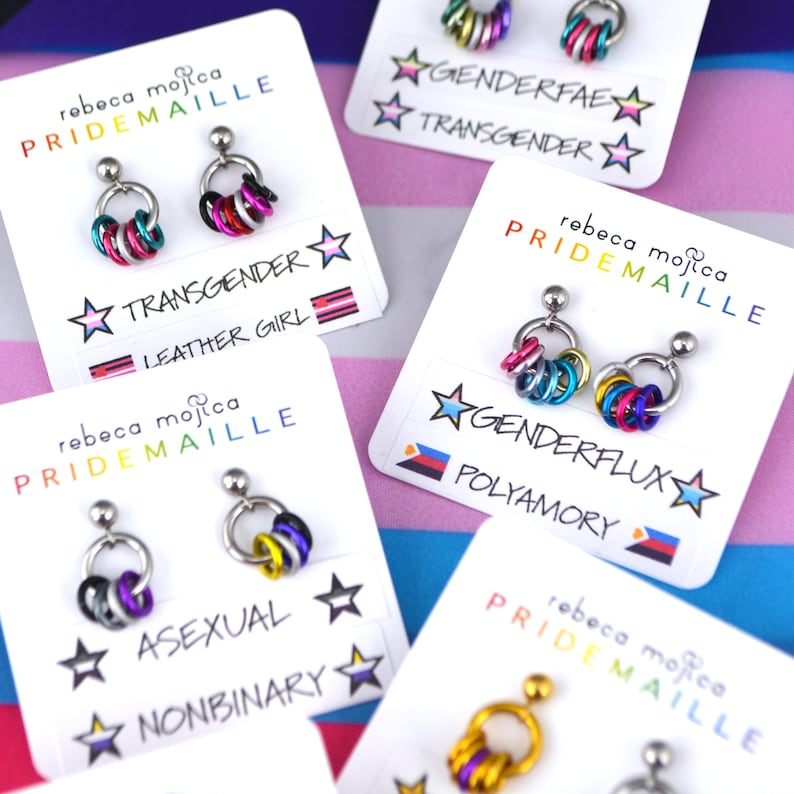 Several pairs of small mismatch pride earrings on cards. Earrings are chainmaille, with one link in each color of the flag. The flags depicted are genderflux, polyamory, asexual, nonbinary, transgender, leather girl, genderfae and intersex