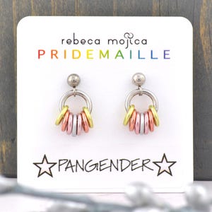 May include: A pair of silver hoop earrings with a cluster of colorful rings hanging from each hoop. The rings are in shades of pink, gold, and silver. The earrings are displayed on a white card with the text "rebeca mojica PRIDEMAILLE" and "PANGENDER" in black.
