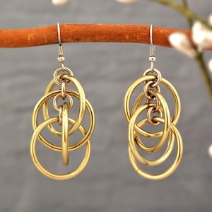 May include: Gold-coloured earrings featuring multiple interlocking circular hoops. The earrings hang from silver-coloured fish hook ear wires. The design showcases a complex arrangement of connected rings, creating a layered and textured appearance.