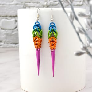 May include: A pair of colourful dangle earrings with a pink spike at the bottom. The earrings are made of metal and have a blue, green, and orange chainmail design.