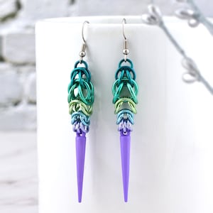 May include: A pair of dangle earrings with a gradient of green, blue, and purple colours. The earrings are made of metal and have a pointed, purple tip.