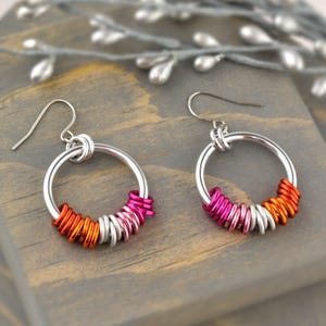 Puede incluir: Un par de pendientes de aro de plata con un diseño en capas de anillos naranja, rosa y plata.
