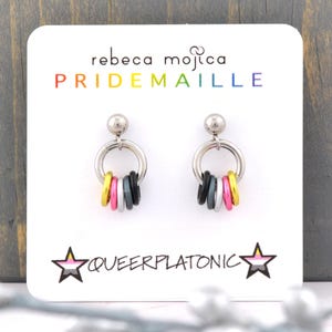 May include: A pair of silver hoop earrings with a charm of five colored rings in the colors of the genderqueer pride flag. The earrings are displayed on a white card with the text "rebeca mojica PRIDEMAILLE" and a star with the text "QUEERPLATONIC" in black and white.