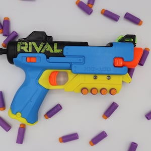 Könnte beinhalten: Ein blauer und gelber Nerf Rival Blaster mit schwarzem Lauf und grünem Visier. Der Blaster ist von lila Schaumstoffpfeilen umgeben.