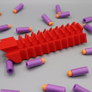 Op de afbeelding: Een rood plastic dartmagazijn met 18 vakjes om darts in te bewaren. Het magazijn is omringd door paarse en oranje foam darts.