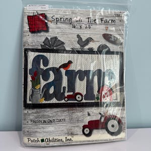 May include: A Spring on the Farm quilt pattern kit in a clear plastic bag. The cover features the text Spring on the Farm 16 x 28, a dark blue farm word graphic with a bird, tractor, and barn, and a red tractor at the bottom. Patch Abilities, Inc.
