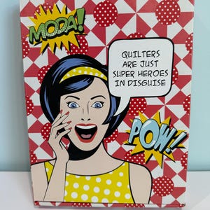 May include: A decorative box with a pop art design. The box features a comic book style illustration of a woman with the text "MODA!" and "POW!" The box also has the text "QUILTERS ARE JUST SUPER HEROES IN DISGUISE". The background is red and white.