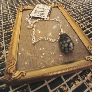 May include: A silver chain necklace with a large, dark stone pendant. The pendant is a speckled black and white stone. The necklace is displayed inside a gold-colored framed glass case. The tag reads "Beverly's Bizarre" and "22 inches".