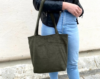 May include: A dark olive green corduroy tote bag featuring a vertical ribbed texture, two matching fabric shoulder straps, and a spacious rectangular body. The bag is constructed from a soft, textured fabric with a consistent deep green tone.