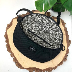 Könnte beinhalten: Schwarze Cordtasche mit Leopardenmuster-Futter. Die Tasche hat einen einzigen Riemen und eine kleine Tasche auf der Vorderseite. Die Tasche liegt auf einer Holzoberfläche.