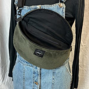 Könnte beinhalten: Olivgrüne Cord-Bauchtasche mit schwarzem Riemen und Reißverschluss. Die Tasche hat ein schwarzes Innenfutter und ein kleines weißes Logo. Getragen über einem Jeans-Latzkleid und einem schwarzen Rollkragenpullover.