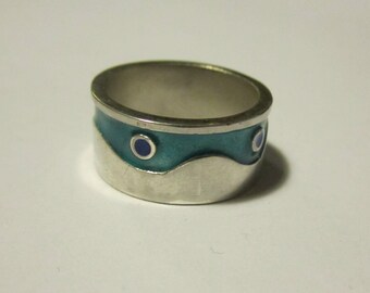 Puede incluir: Un anillo de plata con una banda ancha. El anillo presenta un diseño de esmalte turquesa con un patrón de ondas y dos pequeños círculos azul oscuro. Es una pieza de joyería.
