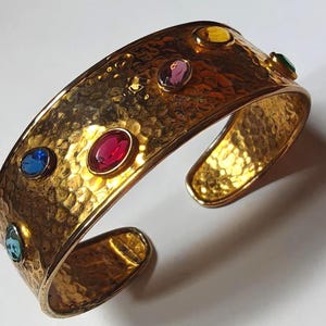 Peut inclure: Un bracelet manchette en métal doré présentant une texture martelée sur toute la surface. Le bracelet est orné de plusieurs pierres facettées de forme ovale et multicolores dans des tons rouge, bleu, bleu clair, jaune et violet.