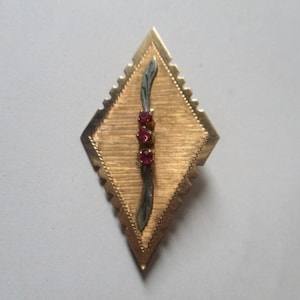 Puede incluir: Un broche dorado en forma de diamante con un diseño de hoja plateada y tres piedras preciosas rojas.