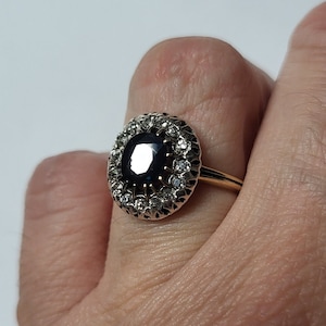 Puede incluir: Un anillo de oro con una gran piedra preciosa azul oscuro rodeada por un halo de piedras preciosas más pequeñas y transparentes.
