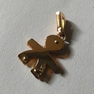 May include: Gold-toned pendant charm in the shape of a stylised person. The charm has a loop at the top for attaching to a necklace or bracelet. The charm is engraved with the word "Gioielli".