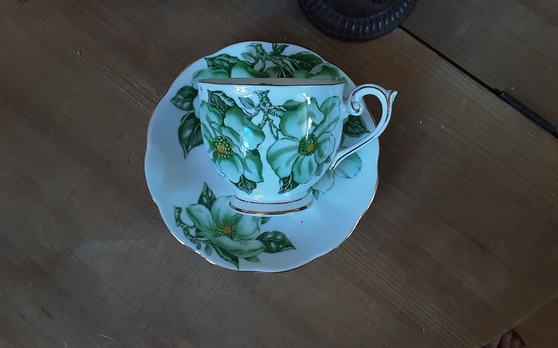 May include: A white teacup and saucer with a green floral design. The cup has a gold rim and the saucer has a scalloped edge.