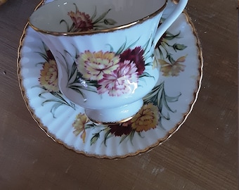 May include: A white teacup and saucer with a gold rim and floral pattern. The flowers are pink, yellow, and red carnations.