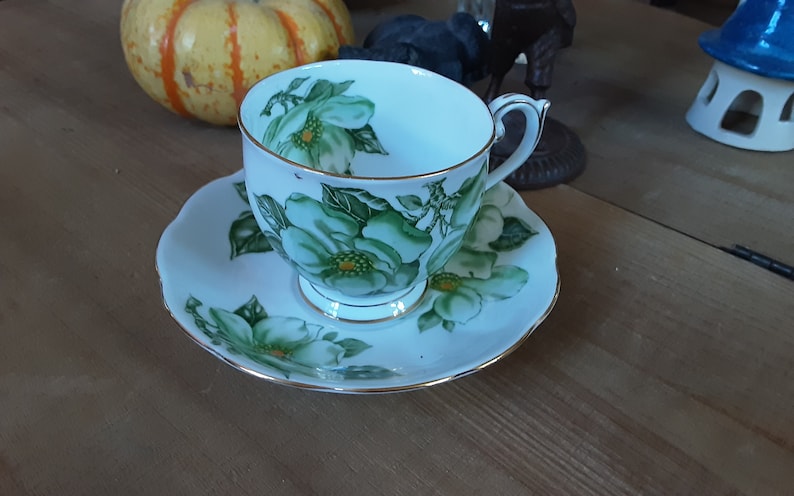 May include: A white teacup and saucer with a green floral design. The cup and saucer have a gold trim.