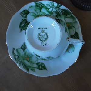 May include: A white teacup and saucer with a green and white floral design. The saucer has a scalloped edge and the teacup has a gold rim. The bottom of the teacup has the words "Magnolia, Queen Anne Fine Bone China England" printed on it.