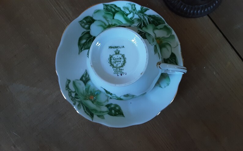 May include: A white teacup and saucer with a green and white floral design. The saucer has a scalloped edge and the teacup has a gold rim. The bottom of the teacup has the words "Magnolia, Queen Anne Fine Bone China England" printed on it.