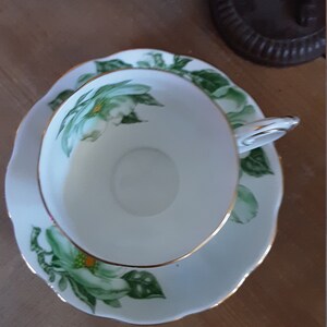 May include: A white teacup and saucer with a green and white floral design. The cup and saucer have a gold trim.