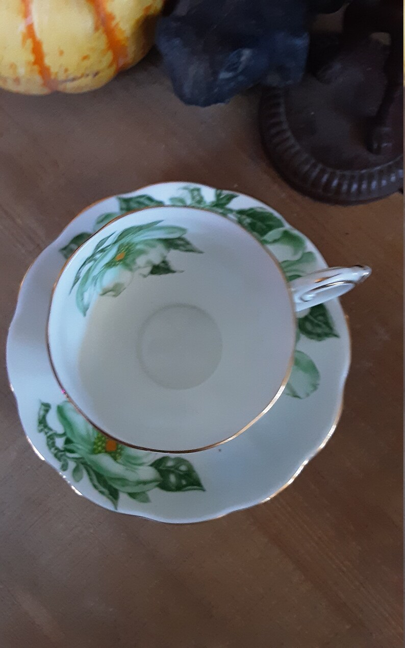 May include: A white teacup and saucer with a green and white floral design. The cup and saucer have a gold trim.