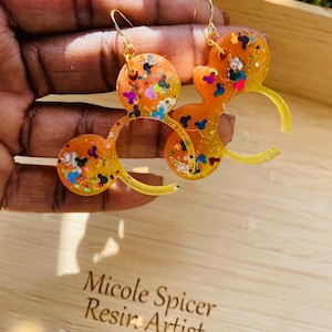 May include: A pair of orange resin earrings shaped like Mickey Mouse ears with colourful glitter embedded in the resin. The earrings are hanging from gold hooks.