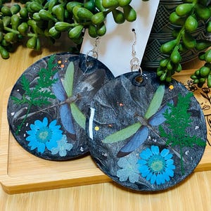 May include: Round earrings with a dark gray background, featuring a dragonfly, blue flowers, and green foliage. The earrings have silver hooks and are displayed on a wooden surface.