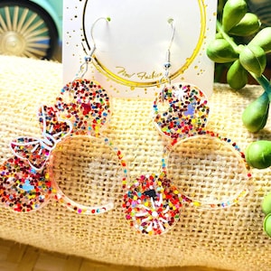 May include: Clear resin earrings with a butterfly and circle design. The earrings are filled with colourful glitter and have silver-coloured hooks. The earrings are displayed on a white card with gold trim.